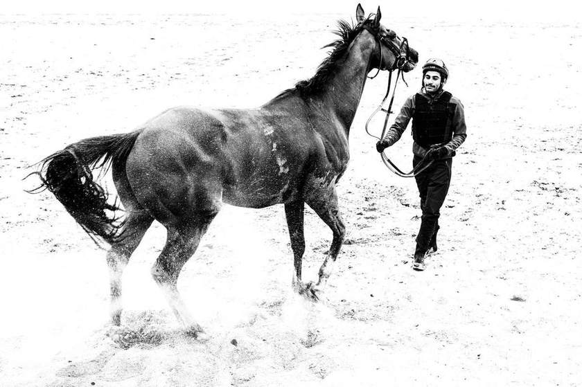 A racehorse being led out on the sand by a member of the yard team, in black and white